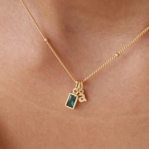 May include: A delicate gold necklace with a small emerald-cut green gemstone pendant and a tiny letter "a" charm. The necklace has a fine chain with small, evenly spaced beads. The jewelry is displayed against a white background.