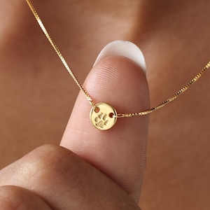 May include: A delicate gold necklace featuring a small, round pendant with a paw print design. The pendant is suspended from a fine chain. The necklace is being held up by a finger, showcasing the jewelry's size and detail.