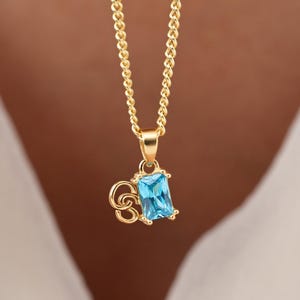 May include: Gold-coloured necklace featuring a rectangular, light blue gemstone pendant. The pendant is suspended from a delicate chain. The necklace is shown against a white background.