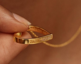 Puede incluir: Un collar con colgante dorado en forma de lágrima. El colgante tiene un diseño de huella dactilar. El collar está sujeto por los dedos de una persona.