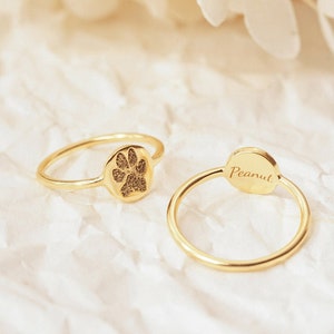 There are oval ring and circle ring were placed on white background. One of the ring has a paw print engraved on the outside, the other one has the name Peanut on the inside of the ring. These gold filled rings are shiny on the background.