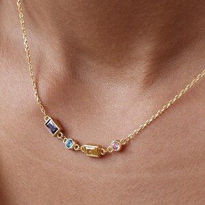 May include: A gold necklace with a delicate chain, featuring a series of colourful gemstones. The necklace has rectangular and round-cut stones in blue, yellow, and pink. The necklace is worn around the neck, showcasing the gemstones.