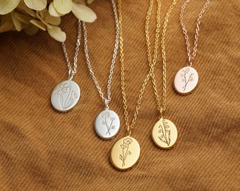 May include: Five necklaces with oval pendants featuring floral designs. The necklaces are in silver, gold, and rose gold tones. The pendants have delicate engraved botanical illustrations. The background is a brown fabric, and dried flowers are in the upper left corner.