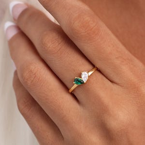 May include: A gold ring featuring two pear-shaped gemstones, one emerald green and one clear, set side by side. The ring is delicate and has a thin band. The gemstones are the focal point of the ring.