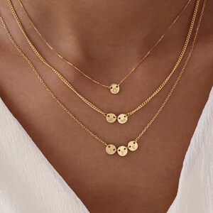 May include: A set of layered gold necklaces. The necklaces have fine chains with small, circular pendants. Some pendants are engraved with initials. The necklaces are worn against a white top.