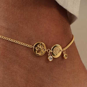 May include: Gold ankle chain with two round charms, one with a paw print and the other with a heart. The chain has two small dangling charms.