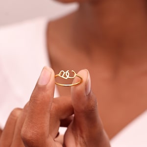 May include: A gold-toned ring featuring two interconnected letters, B and C, as the central design element. The ring has a thin, smooth band and a polished finish, showcasing the stylized, open-work lettering that sits atop the circular band.