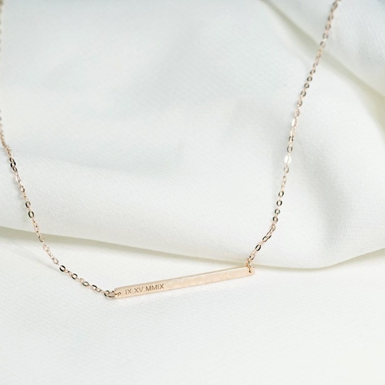 May include: A delicate gold chain necklace with a thin, flat bar pendant engraved with the Roman numerals "IX XV MMXIX".