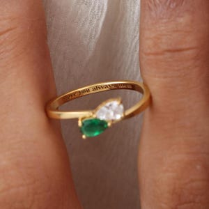 May include: A gold ring with a pear-shaped emerald and a white gemstone. The ring has the inscription "love you always, mom" engraved on the band. The ring is on a finger.