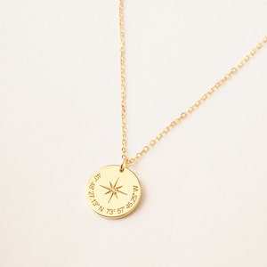 A gold circle necklace is engraved with a compass symbol, and a coordinates underneath it. The chain is set to make a curve line on the grey background. The gold chain is shiny, and the gold circle pendant is outstanding on the grey background.