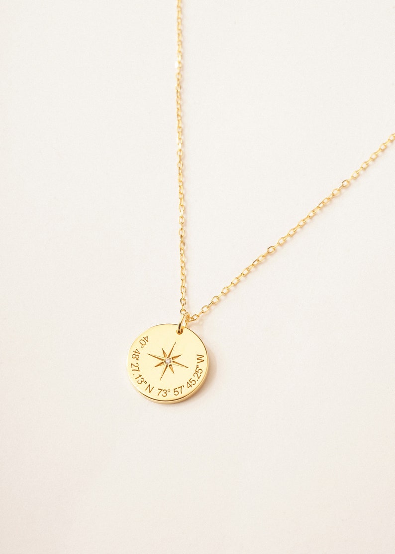 A gold circle necklace is engraved with a compass symbol, and a coordinates underneath it. The chain is set to make a curve line on the grey background. The gold chain is shiny, and the gold circle pendant is outstanding on the grey background.