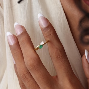 May include: A gold ring featuring two pear-shaped gemstones, one white and one emerald green. The ring is worn on a finger, showcasing its delicate design and vibrant colors. The ring's band is thin and elegant.