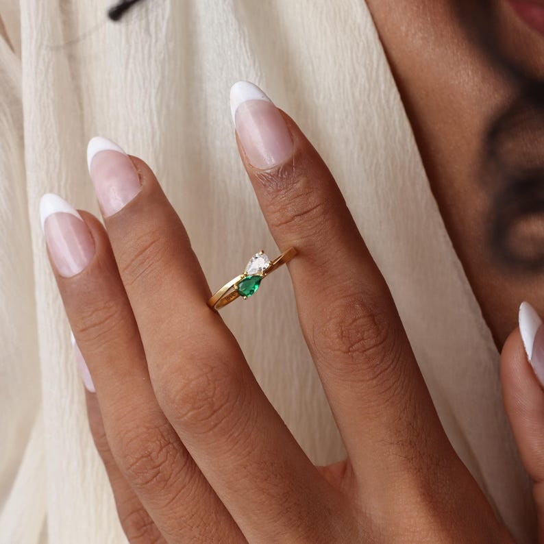 May include: A gold ring featuring two pear-shaped gemstones, one white and one emerald green. The ring is worn on a finger, showcasing its delicate design and vibrant colors. The ring's band is thin and elegant.
