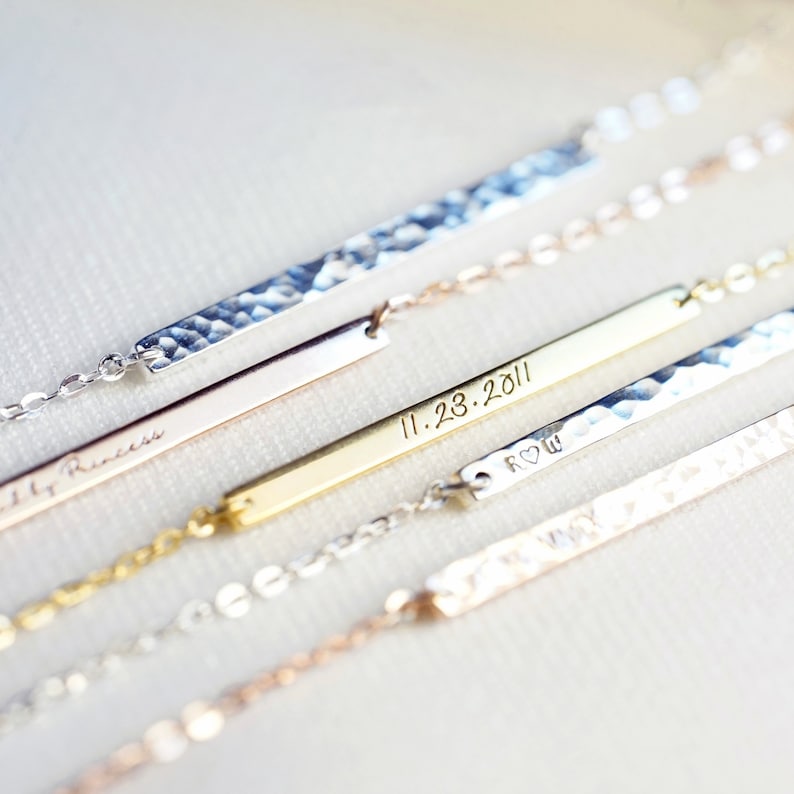 May include: Four delicate gold bar necklaces with different engraved messages. The necklaces feature a hammered texture and are layered on a white surface.