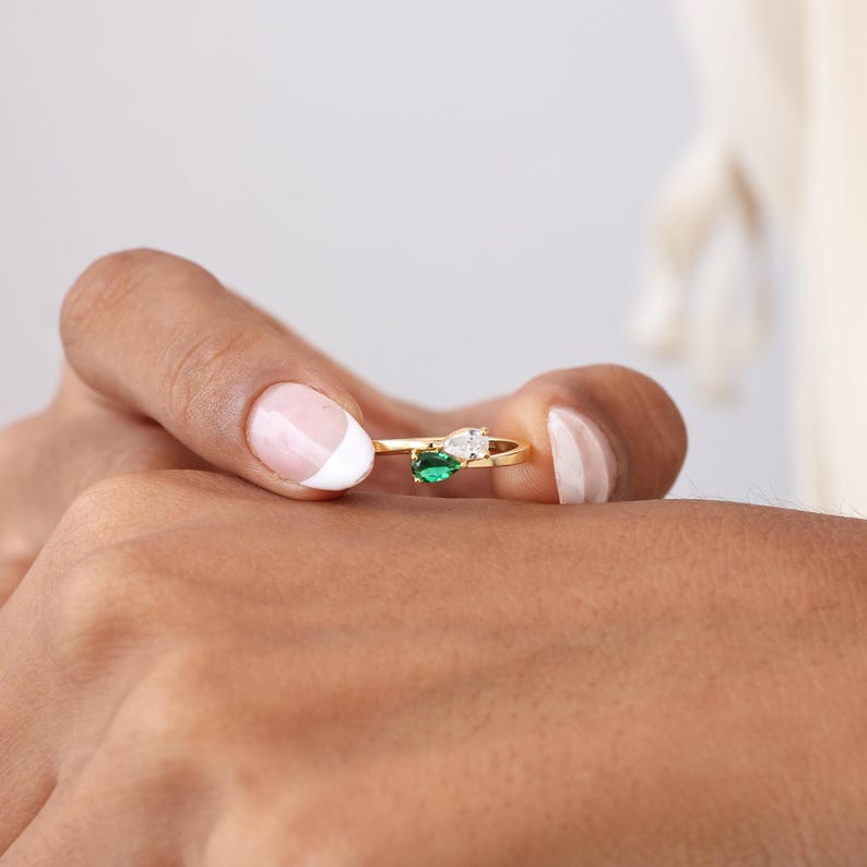 May include: A gold ring featuring an oval green gemstone and a pear-shaped white gemstone. The ring is held between two fingers, showcasing the delicate design and the sparkle of the gemstones.