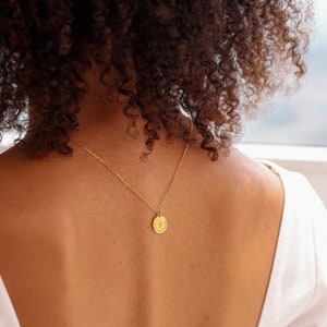 May include: A gold-toned necklace featuring a small, circular pendant with an engraved starburst design. The pendant hangs from a delicate, thin gold-toned chain. The circular charm is centered on the chain, resting against the upper back area.