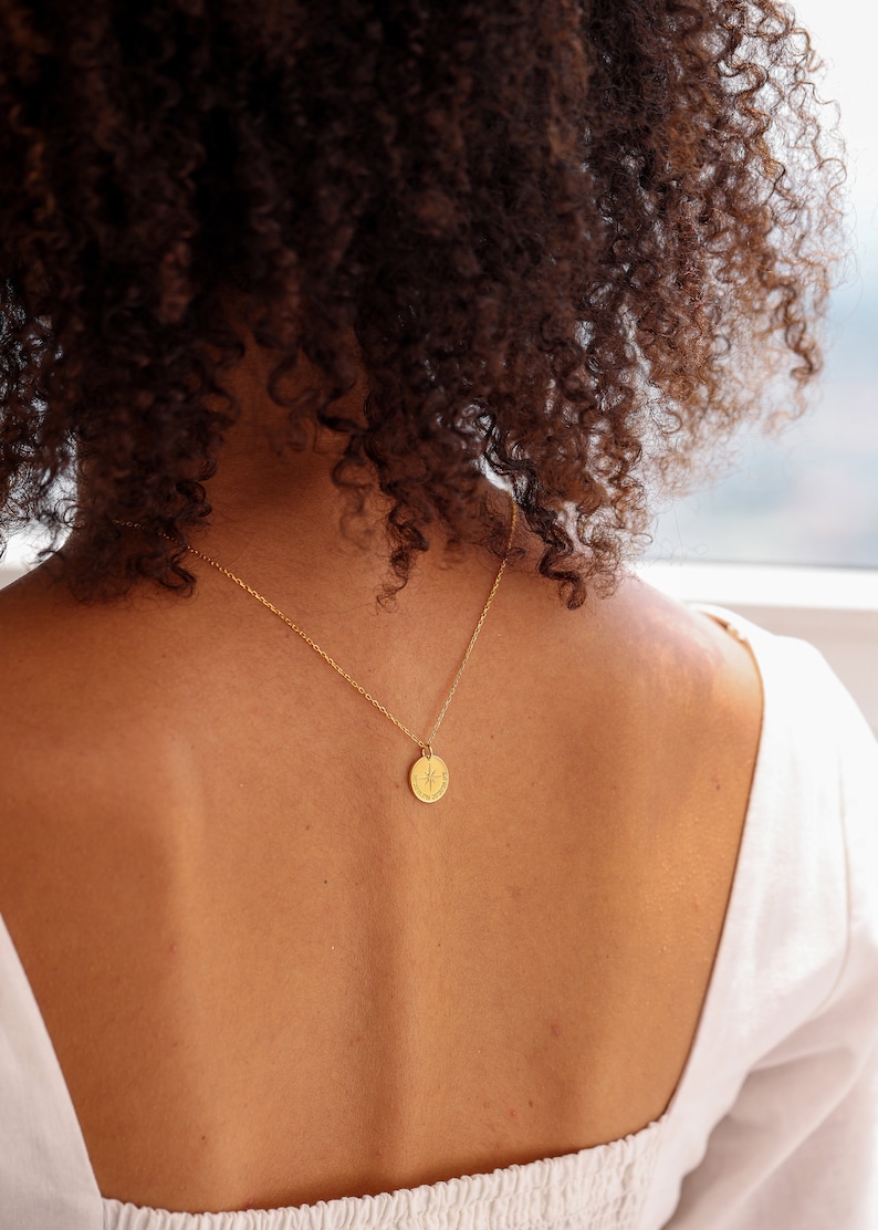 May include: A gold-toned necklace featuring a small, circular pendant with an engraved starburst design. The pendant hangs from a delicate, thin gold-toned chain. The circular charm is centered on the chain, resting against the upper back area.