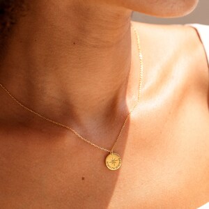 May include: A gold-toned necklace featuring a small, circular pendant with an engraved compass rose design and the coordinates 36&deg;20'11.7"N 25&deg;26'28.9"E. The pendant hangs from a delicate, thin gold-toned chain that rests against the collarbone area.