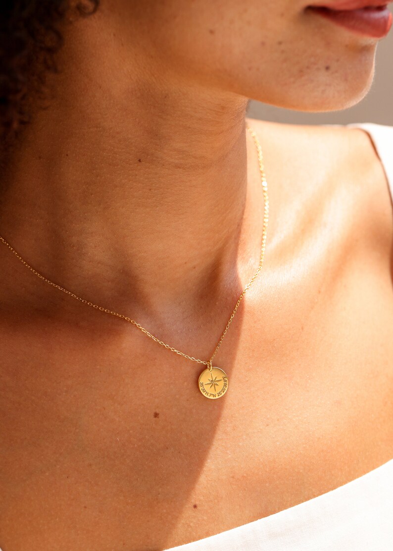 May include: A gold-toned necklace featuring a small, circular pendant with an engraved compass rose design and the coordinates 36&deg;20'11.7"N 25&deg;26'28.9"E. The pendant hangs from a delicate, thin gold-toned chain that rests against the collarbone area.