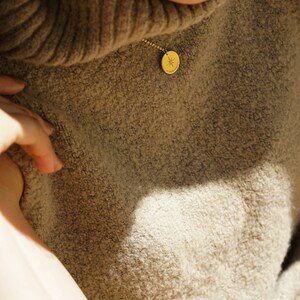 May include: A gold-toned circular pendant necklace featuring a delicate chain and a small round charm engraved with a starburst design. The pendant rests against a textured, beige-colored knit sweater with a high collar, capturing light.