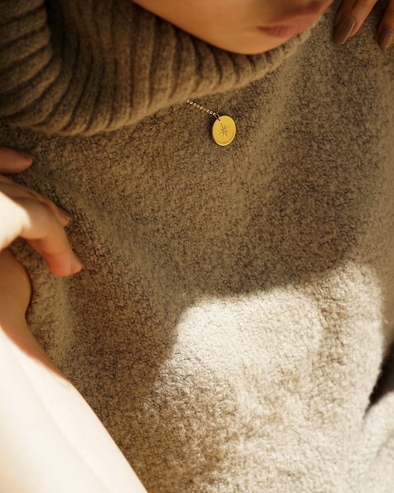 May include: A gold-toned circular pendant necklace featuring a delicate chain and a small round charm engraved with a starburst design. The pendant rests against a textured, beige-colored knit sweater with a high collar, capturing light.