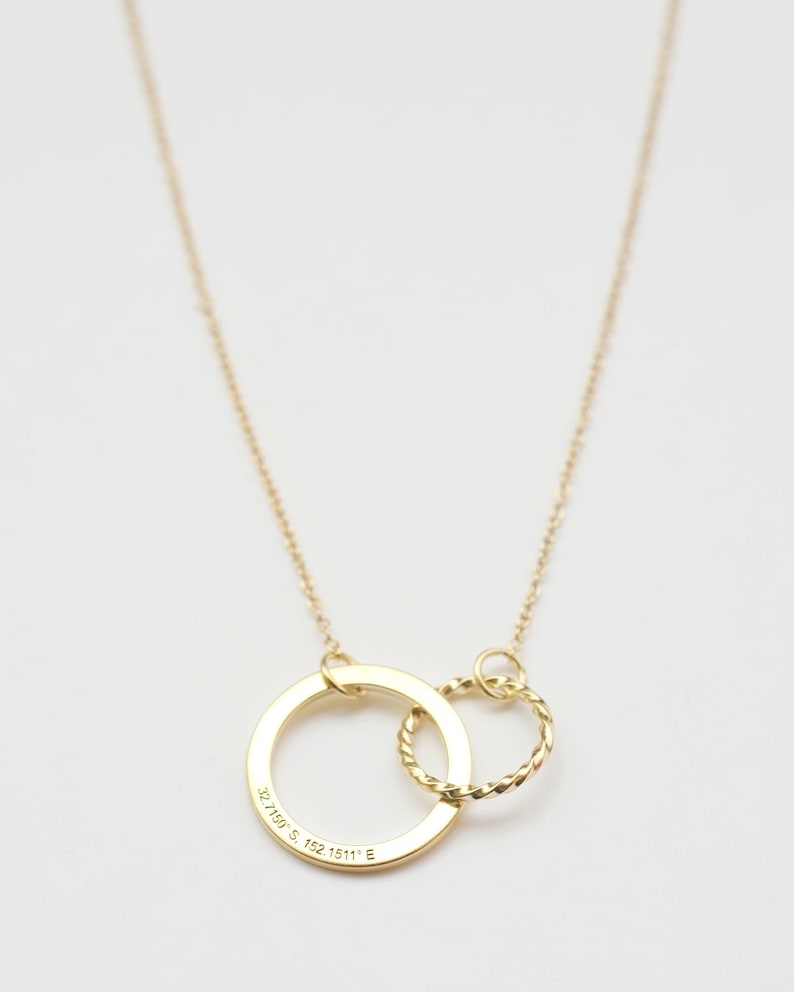 May include: A gold necklace with two interlocking circles. The larger circle has the coordinates "32.7489&deg; S, 152.1511&deg; E" engraved on it.