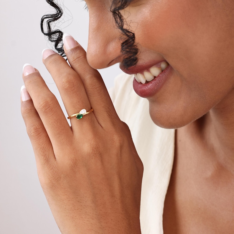 May include: A gold ring featuring a small, heart-shaped green gemstone and a white gemstone. The ring is on a person's finger, with the person's hand and face visible in the image.
