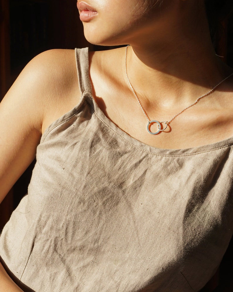 May include: A silver necklace with two interlocking circles hangs on a person wearing a light brown tank top.