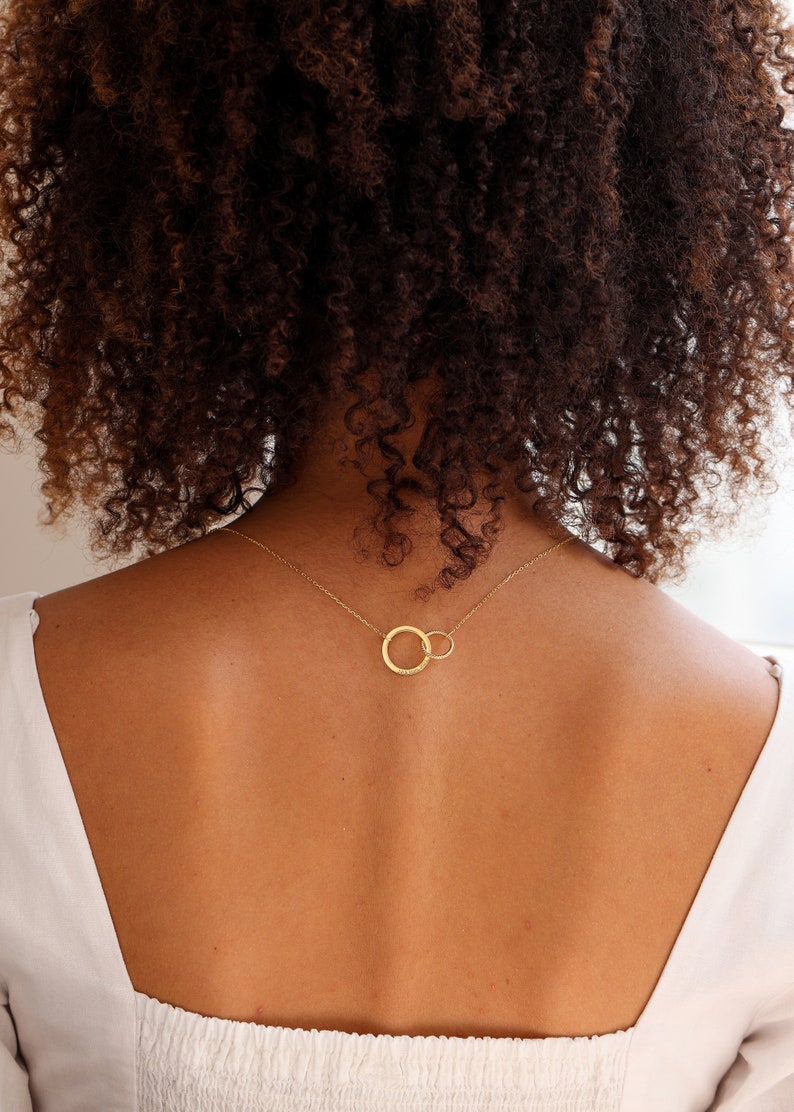 May include: A gold necklace with two interlocking circles. The necklace is worn by a person with curly brown hair.