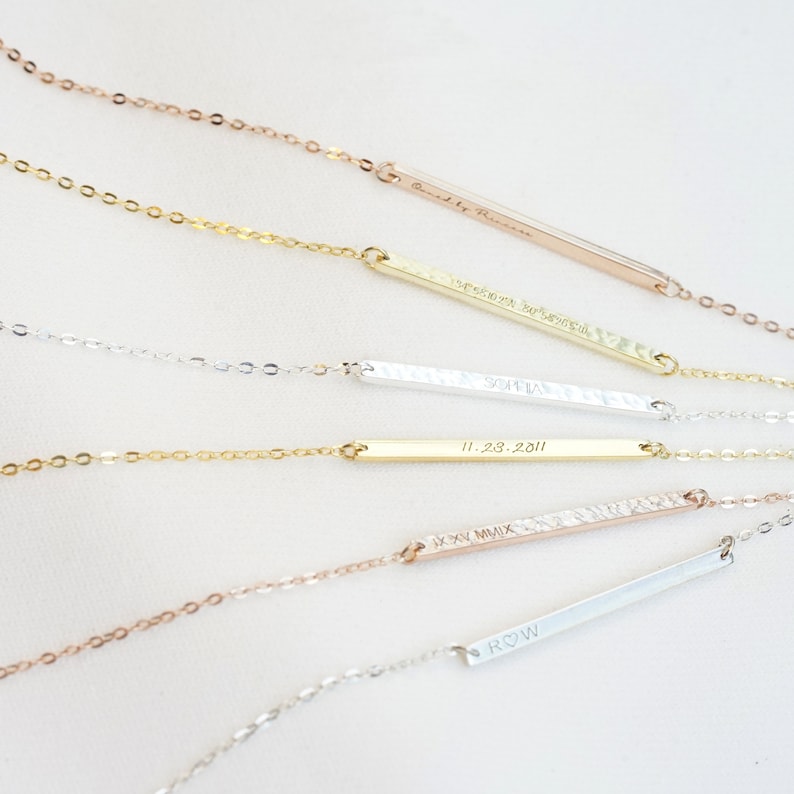 May include: A collection of personalized bar necklaces with different engravings. The necklaces are made of gold, silver, and rose gold chains with matching bar pendants. The engravings include names, dates, and Roman numerals.