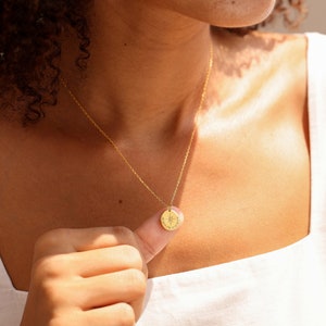 May include: A gold-toned necklace featuring a delicate chain and a small, circular pendant. The pendant is engraved with a central star-like compass design and small text along the bottom edge, suspended from a thin, polished metal link chain.