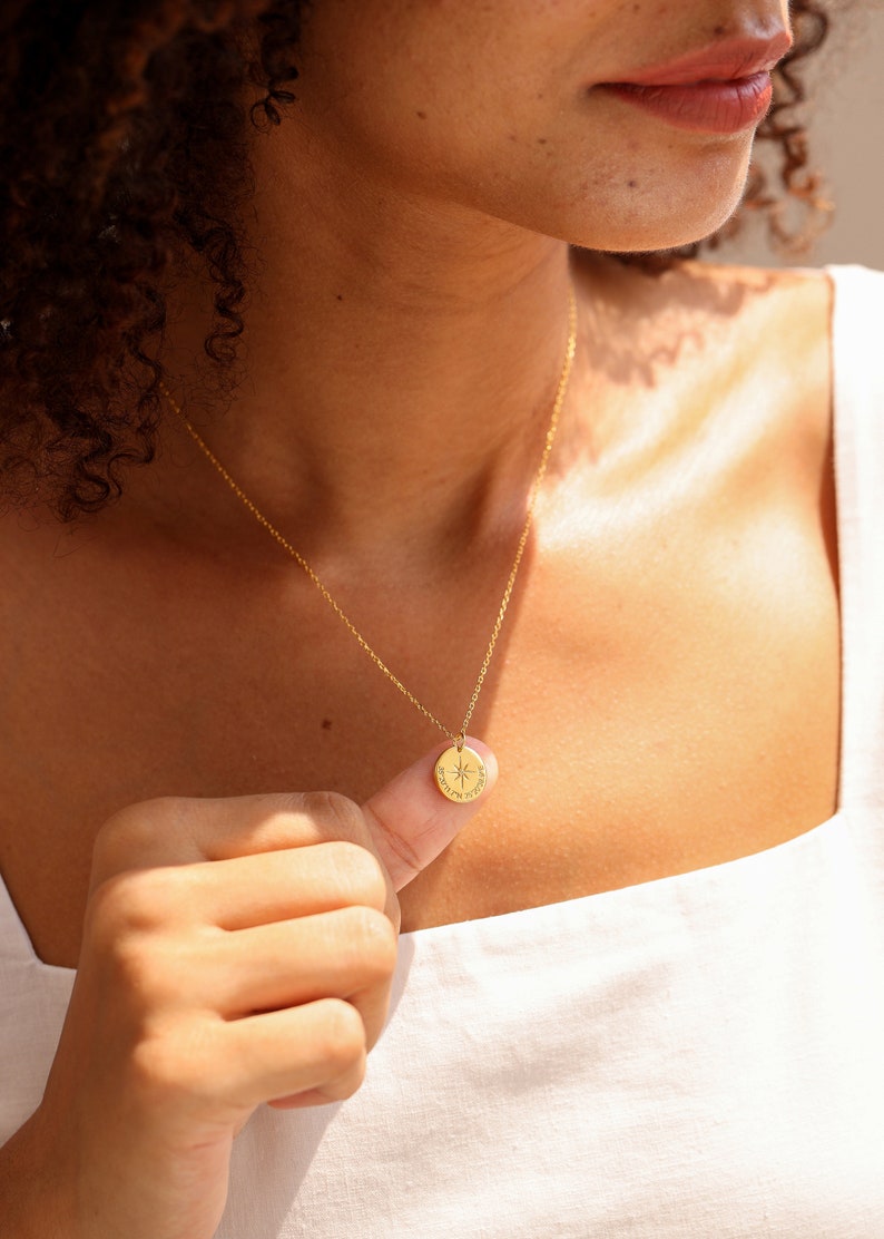 May include: A gold-toned necklace featuring a delicate chain and a small, circular pendant. The pendant is engraved with a central star-like compass design and small text along the bottom edge, suspended from a thin, polished metal link chain.