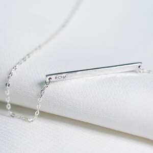 May include: A silver necklace with a thin, hammered bar pendant. The pendant is engraved with the letters "R" and "W" with a heart symbol between them.