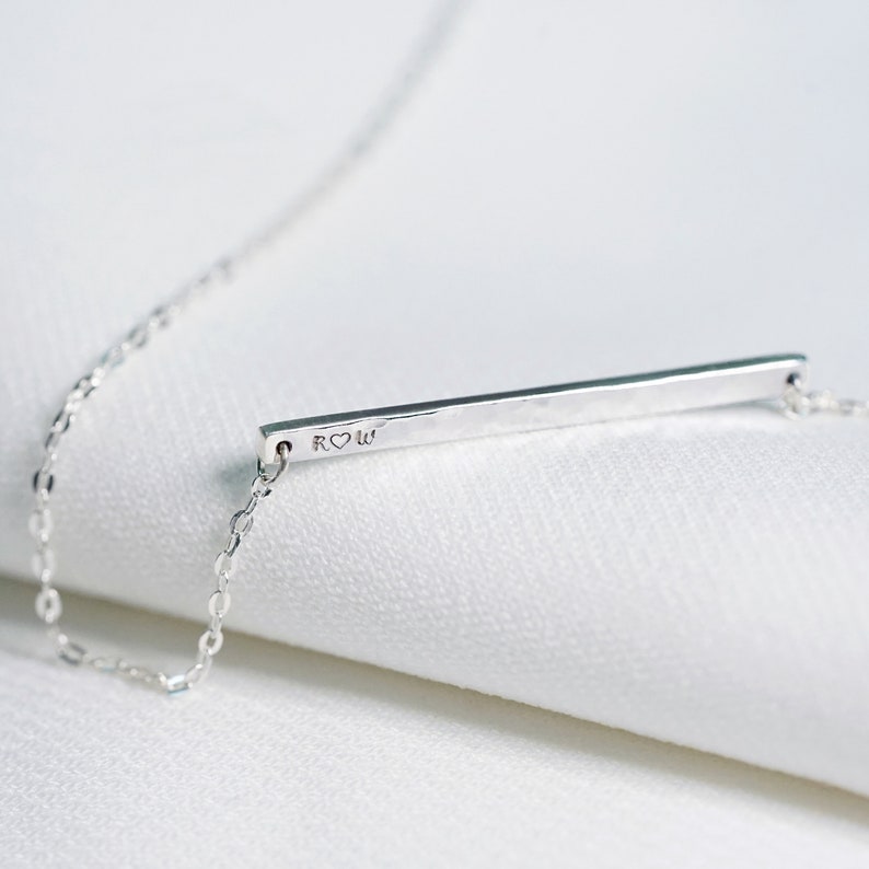 May include: A silver necklace with a thin, hammered bar pendant. The pendant is engraved with the letters "R" and "W" with a heart symbol between them.