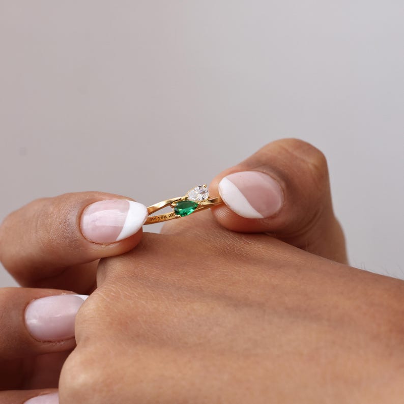 May include: A gold ring featuring a pear-shaped emerald and a round, clear gemstone. The ring has a delicate band with the inscription "love you always". The ring is held between two fingers.