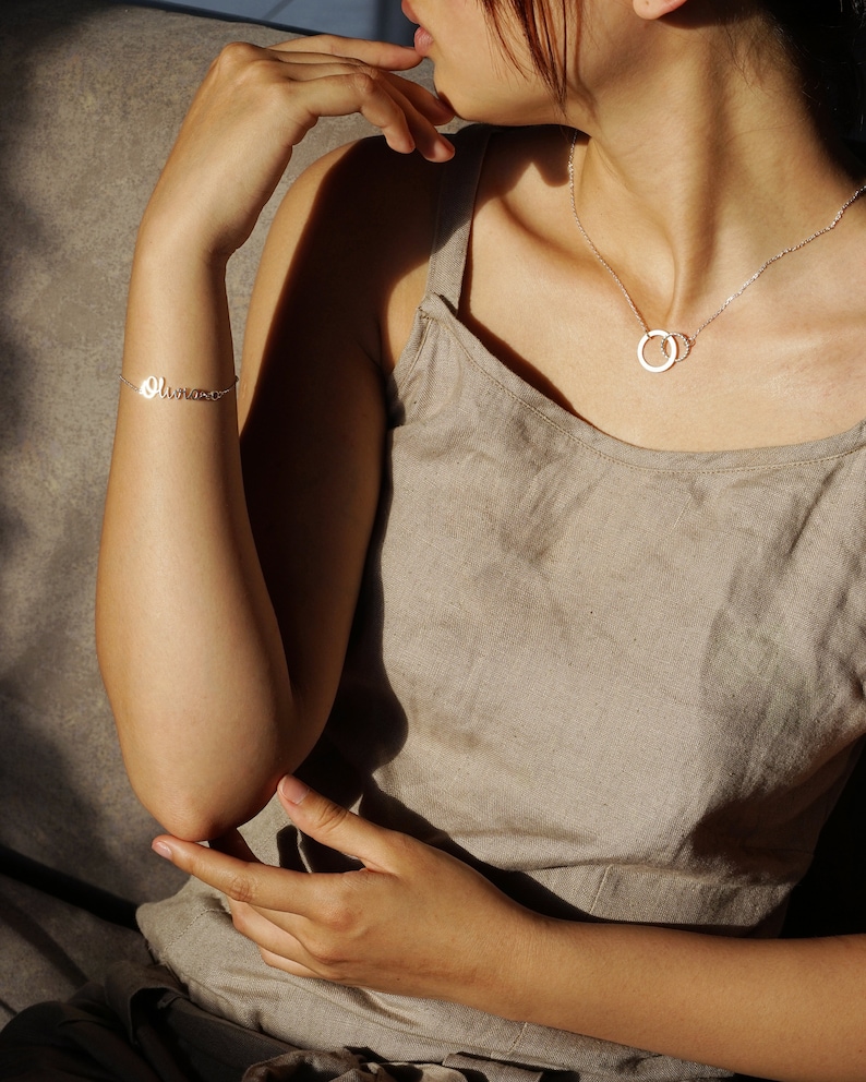 May include: A woman wearing a simple, light brown linen tank top with a silver necklace featuring two interlocking circles. She is also wearing a silver bracelet with the word "Olivia" inscribed on it.