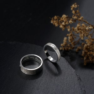A couple silver ring is standing on a black stone. One ring for men, which is 7mm wide, and one ring for woman, which is 4mm wide. The fingerprint is engraved outside of the ring, and the inscription is engraved inside of the ring.