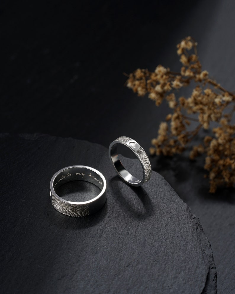 A couple silver ring is standing on a black stone. One ring for men, which is 7mm wide, and one ring for woman, which is 4mm wide. The fingerprint is engraved outside of the ring, and the inscription is engraved inside of the ring.