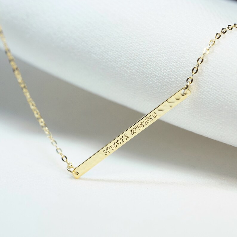 May include: A gold necklace with a thin bar pendant engraved with the coordinates "34&deg; 58'10.2"N 80&deg; 58'28.5"W".