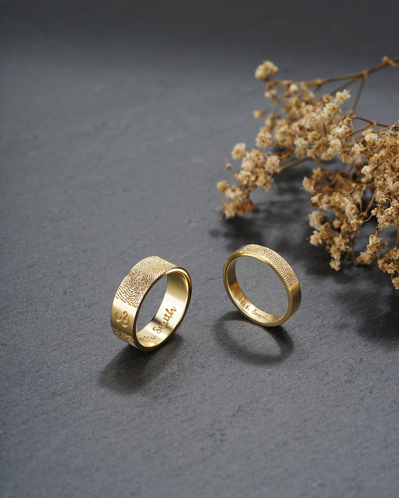 A couple gold ring is standing on a black stone. One ring for men, which is 7mm wide, and one ring for woman, which is 4mm wide. The fingerprint is engraved outside of the ring, and the inscription is engraved inside of the ring.