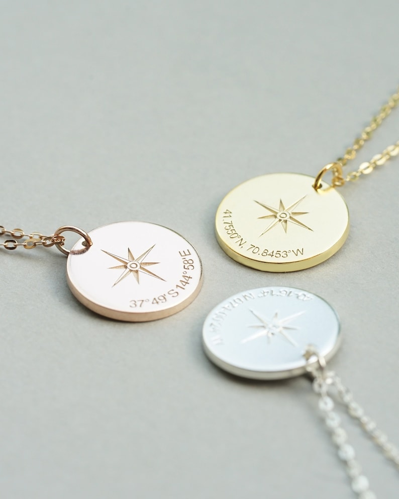 May include: Three circular pendant necklaces in gold, rose gold, and silver tones. Each pendant features an engraved eight-pointed compass star at the center and geographic coordinates etched along the bottom edge of the circular metal surface.