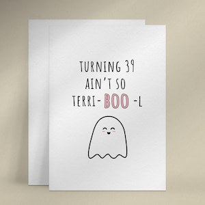 Puede incluir: Una tarjeta de felicitación blanca con el texto "TURNING 39 AIN'T SO TERRI-BOO-L" en letras negras y rosas. Debajo del texto hay un fantasma de dibujos animados con una cara sonriente. La tarjeta va acompañada de un sobre blanco.
