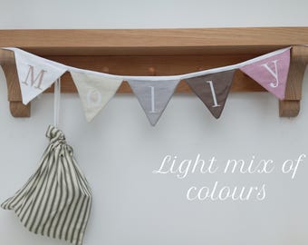 Op de afbeelding: Een houten plank met een slinger met de naam "Molly" op driehoekige vlaggen in wit, beige, grijs, bruin en roze. Een gestreepte trekkoordtas hangt eronder. Tekst: "Light mix of colours".