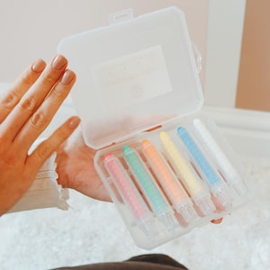 May include: A clear plastic case with a label that says "Crayola Crayons" holds seven colourful crayons. The crayons are arranged in a row and are in the colours red, orange, yellow, green, blue, light blue, and white.