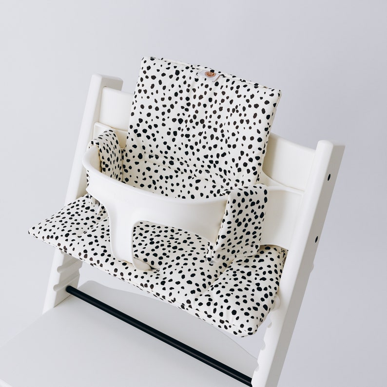 May include: White high chair with a black and white spotted cushion. The cushion is made of fabric and has a soft, comfortable feel. The cushion is designed to fit snugly in the high chair and provides extra support for the child.