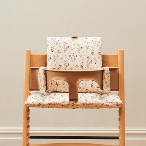 May include: A wooden high chair with a beige seat and back cushion. The cushions are patterned with small brown floral designs. The chair has a beige safety bar and a footrest. The chair is designed for a baby or toddler.