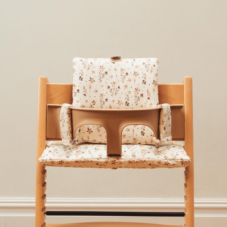 May include: A wooden high chair with a beige seat and back cushion. The cushions are patterned with small brown floral designs. The chair has a beige safety bar and a footrest. The chair is designed for a baby or toddler.