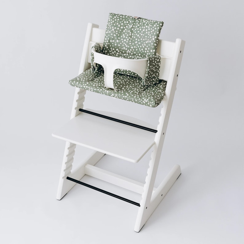 Op de afbeelding: Witte hoge stoel met een groen-wit gepatroneerd kussen en een wit dienblad. De stoel heeft een zwarte metalen balk aan de voorkant.
