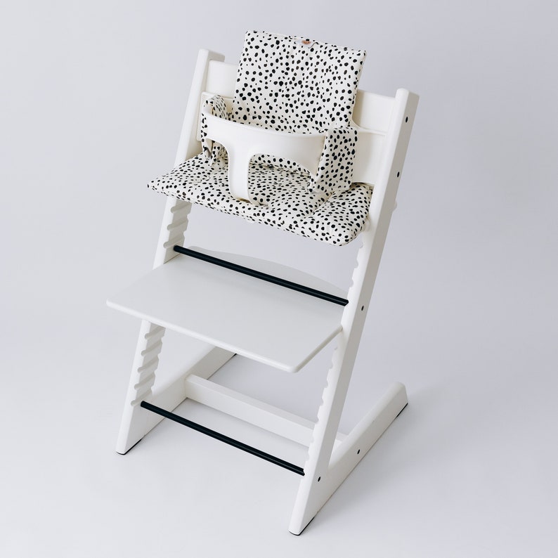May include: White high chair with a black and white polka dot seat cushion. The chair has a tray and a safety harness.