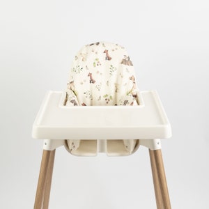 Puede incluir: Una trona con asiento y bandeja color crema, y patas de madera. La funda del asiento presenta un estampado de criaturas del bosque como zorros y osos, junto con hojas y mariposas, sobre un fondo crema. Para beb&eacute;s y ni&ntilde;os peque&ntilde;os.